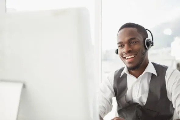 Carrera Software | A smiling man wearing a headset and vest sits at a desk, looking at a computer screen in a bright office setting, exploring a teamviewer alternative for remote support.