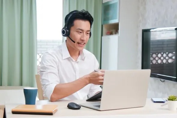 Carrera Software | A man wearing a headset sits at a desk, smiling and pointing at his laptop during a video call. In the bright room, he discusses the best TeamViewer alternative with colleagues, surrounded by a coffee cup, notebook, and mouse.