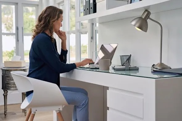 Carrera Software | A woman sits at a modern white desk, working on a laptop in a bright, organized home office. Focused with her hand on her chin, she appears to be searching for a teamviewer alternative among office supplies and a desk lamp.